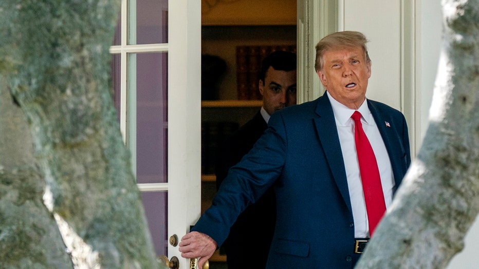 Trump, wearing blue suit and red tie, holds Oval Office door; an aide is seen behind him
