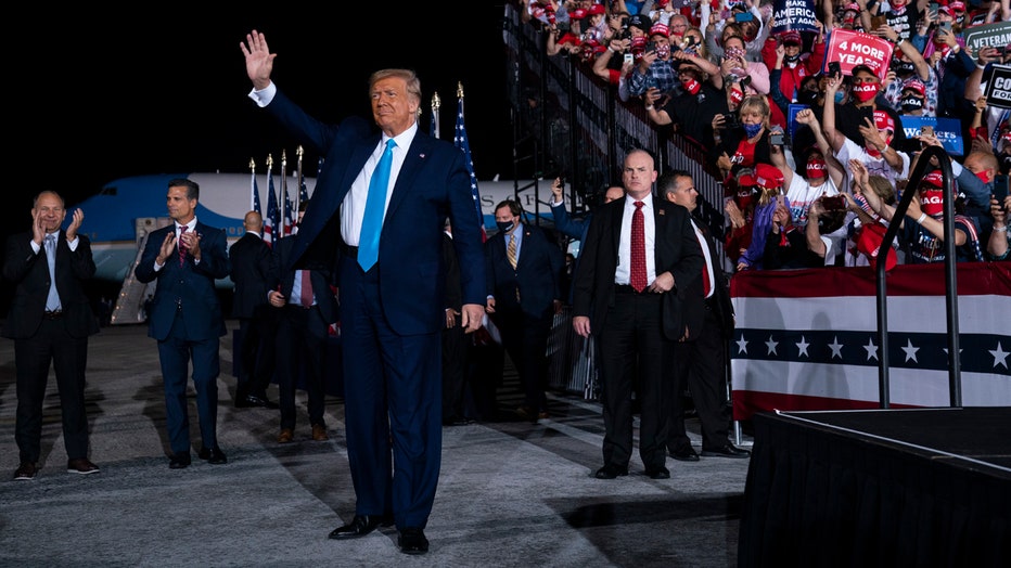 Donald Trump waves at a rally; in the background are supporters in bleachers, security personnel, staff, American flags, Air Force One