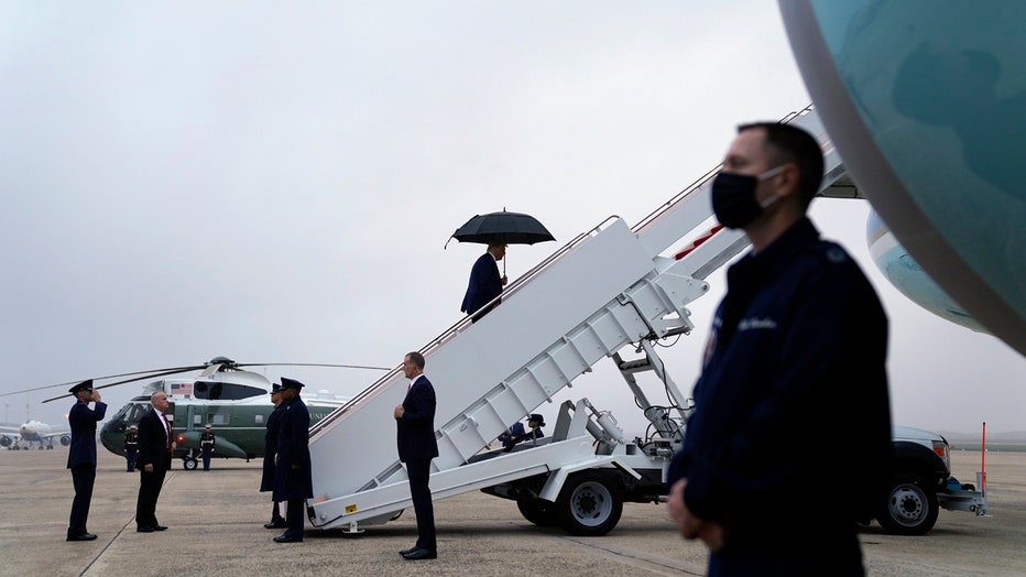 Trump, holding an umbrella, walks up mobile stairs to board Air Force One; a member of the military salutes as other military members and staffers stand on the tarmac