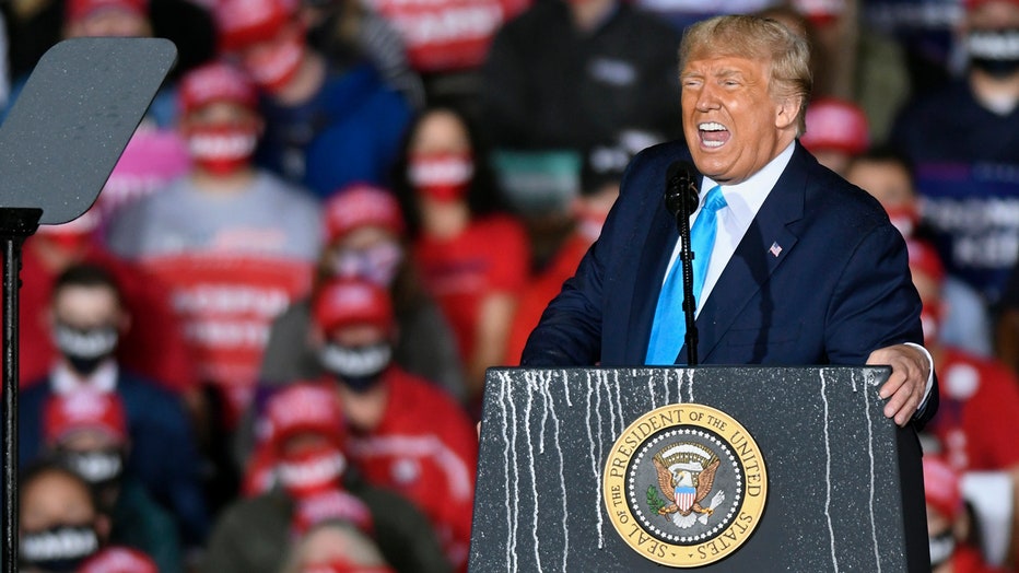 Trump, wearing a dark blue suit and a light blue tie, speaks from behind a lectern featuring the presidential seal; supporters are in the background