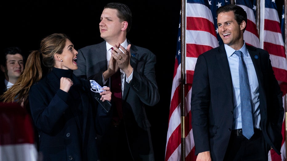Hope Hicks, holding a mask, cheers and makes a fist gesture at a rally; three men and some American flags are visible