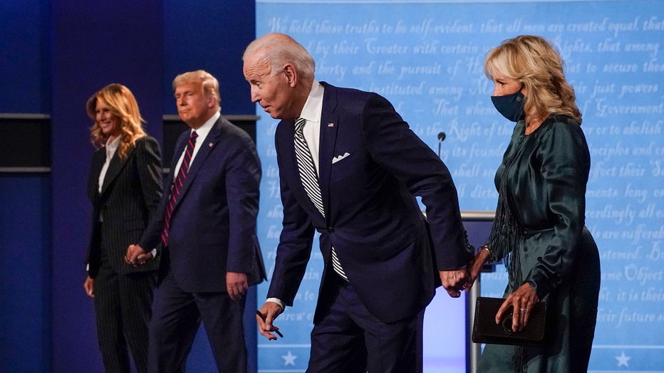 Melania Trump, wearing a striped suit, and Donald Trump, wearing a blue suit and a blue-and-red tie, hold hands; Joe Biden, wearing a blue suit and a blue-and-white tie, and Jill Biden, wearing a sage-green outfit, hold hands