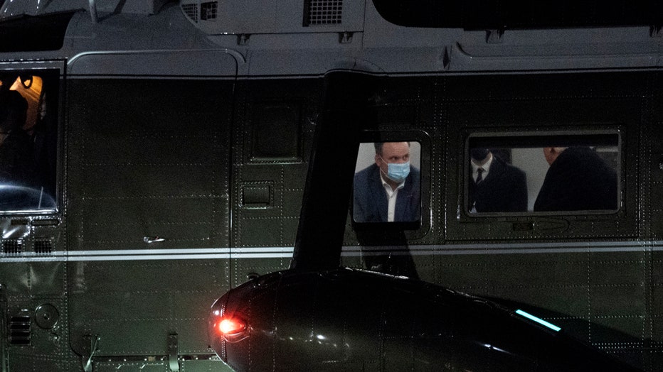 Three people are seen through windows of the Marine One helicopter; one of the people, Dan Scavino, is wearing a light blue mask