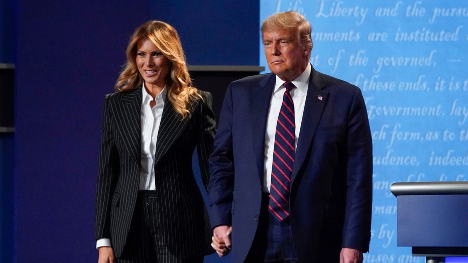 Melania Trump, wearing a striped suit, smiles as she holds Donald Trump's hand on stage; the president is wearing a blue suit and a blue-and-red tie