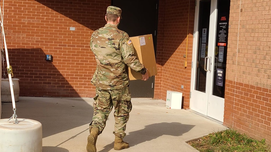 Military member in camo carries a box towards the door of a clinic