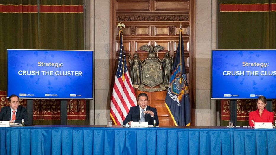 Cuomo and two others sit behind a long table with flags and screens behind them