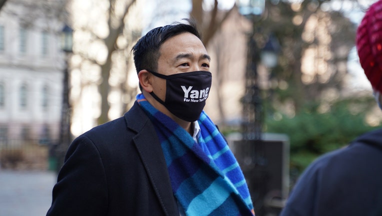 Andrew Yang wearing a black coat, blue patterned scarf, and black face mask with the word 'Yang', standing outdoors with City Hall in background