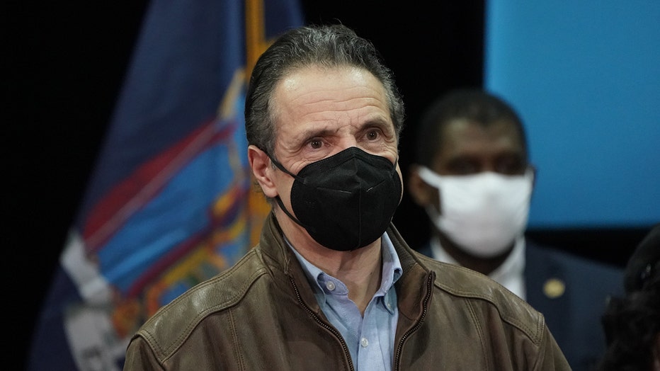 Andrew Cuomo wearing a black mask, light blue shirt, and brown leather jacket