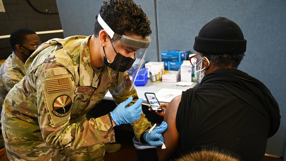 Air Force member in camo and face shield gives a shot in the arm of a seated person