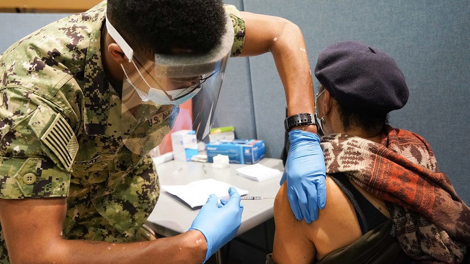 Navy sailor wearing a face shield and mask and camouflage uniform gives an injection to a seated person