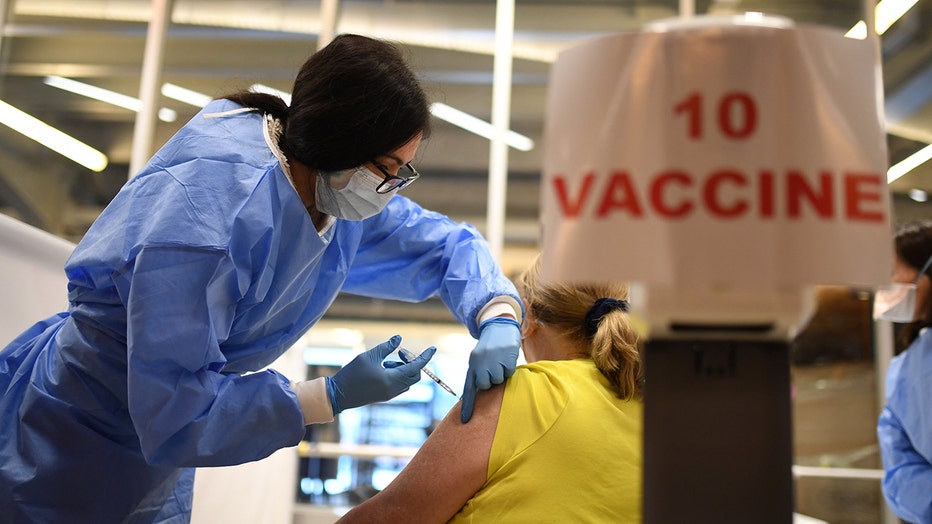Medical worker in light blue gown and mask gives a shot to a woman whose back is to camera