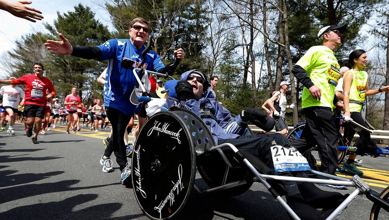 An athlete in a blue shirt and sunglasses holds out his hand for a high-five as he pushes his son in a special racing wheelchair. They are among dozens of runners on a tree-lined road in the Boston Marathon on a sunny day.