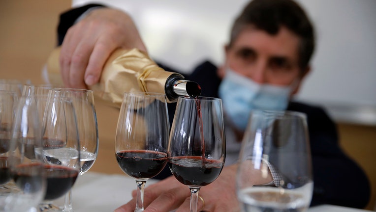 A man wearing a mask pours wine from a wrapped bottle into wine glasses