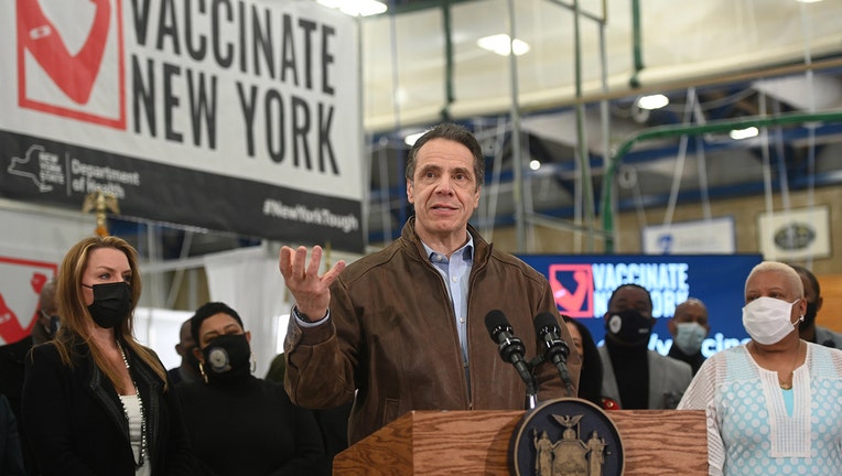 Cuomo in a brown jacket speaks from behind a lectern as other people stand near him; a large Vaccinate New York banner hangs from the ceiling