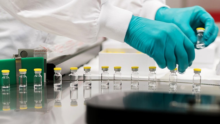 Close-up of a row of small glass vials with yellow caps. A person's hands in green gloves holds two vials.
