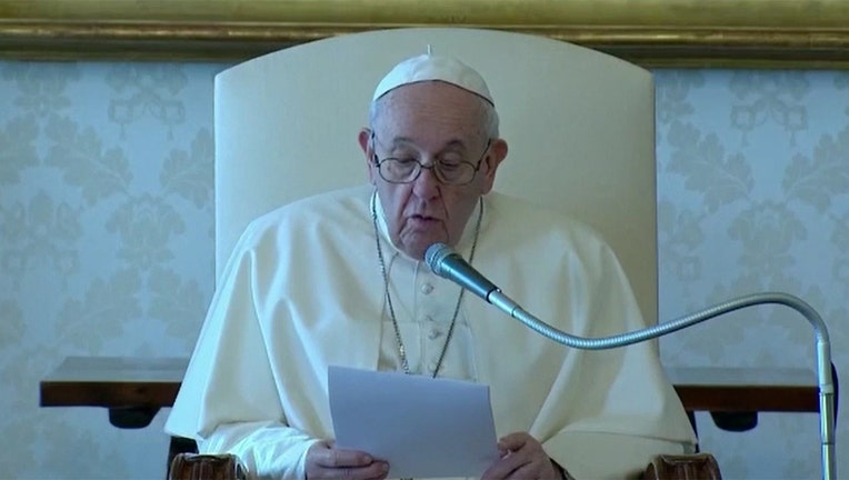 Pope Francis wearing white skullcap and vestments and glasses holds a document and speaks into a microphone