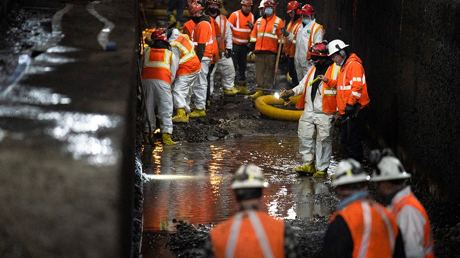 More than a dozen workers in hard hats and safety gear work inside a partially flooded rail tunnel; two workers shine flashlights at the ground