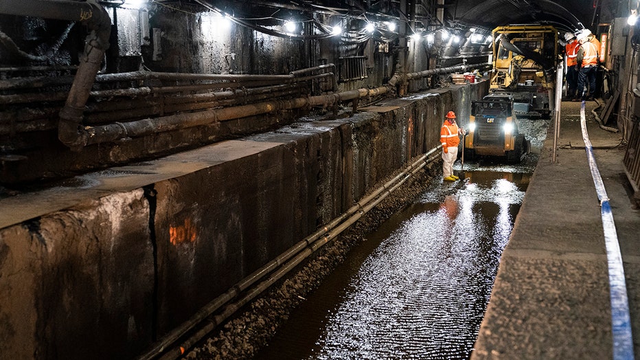 Workers in orange safety gear stand near heavy construiction vehicles inside a partially flooded rail tunnel