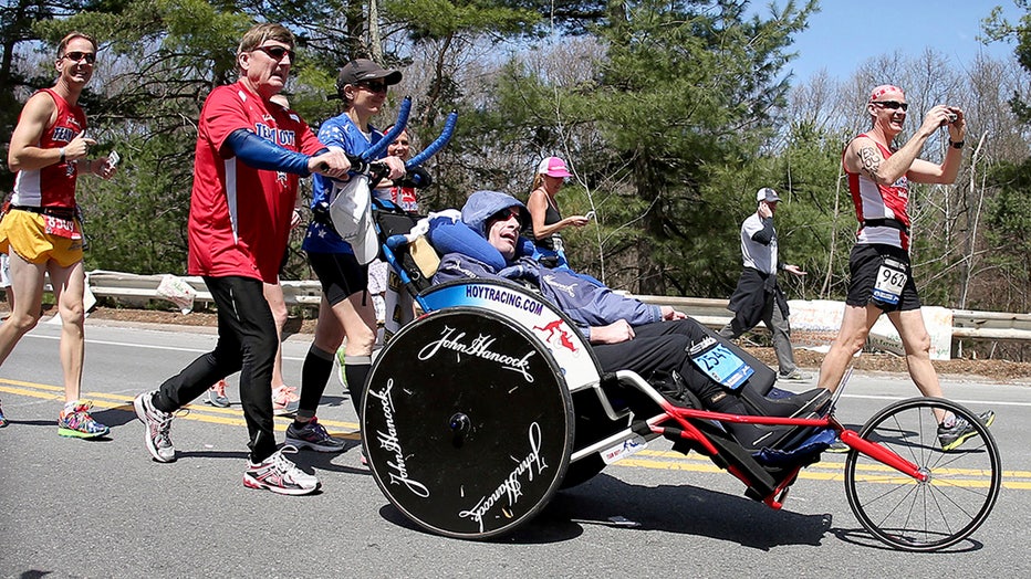A man in a red shirt, black track pants and sunglasses pushes his son in a racing wheelchair along a suburban road during the Boston Marathon. Several runners are around them.
