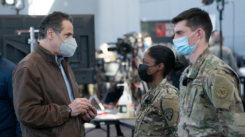 Cuomo wearing mask and brown jacket speaks to two service members wearing masks and camouflage uniforms