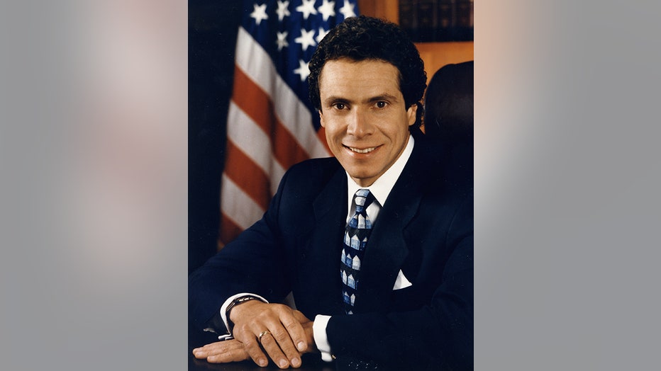 Andrew Cuomo in blue suit and tie smiles with hands crossed on table in front of him and US flag behind him