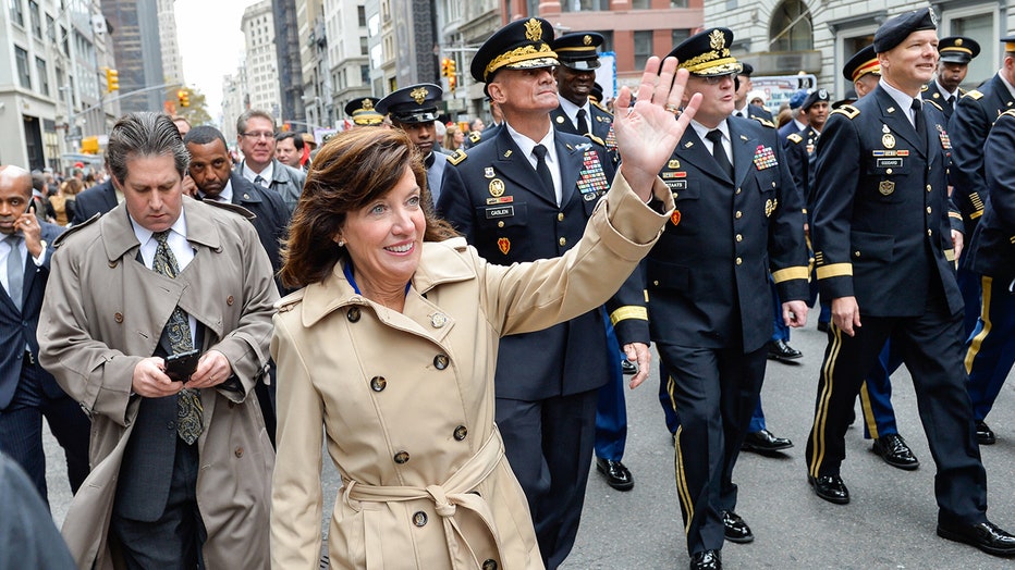 Kathy Hochul in a tan trench coat smiles and waves as she marches in front of uniformed soldiers