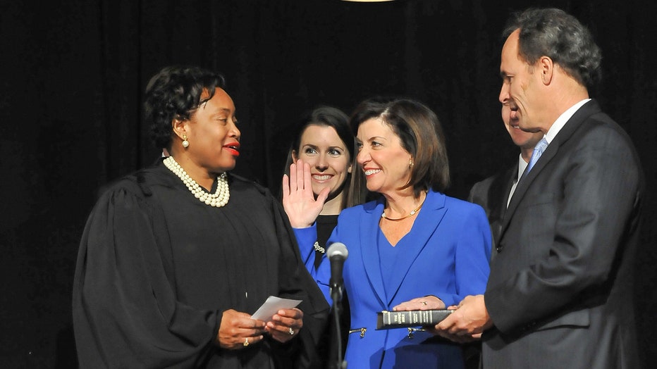 A smiling Kathy Hochul in a blue suit holds right hand up and left hand on a bible as judge administers the oath of office; a man stands to her left and a woman stands behind her