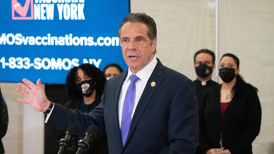 Gov. Cuomo in a dark suit and purple tie speaks at a vaccine site and holds his right arm and hand up away from his body