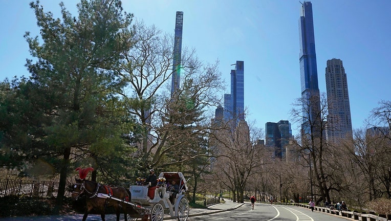 A horse and carriage make progress on a road inside Central Park in New York; very tall and narrow luxury towers rise into a blue sky