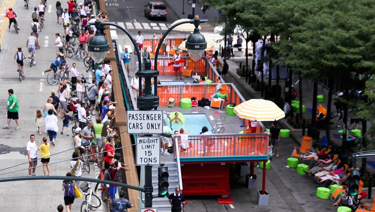 Small plunge pools built inside dumpsters set up on Park Avenue in New York; some people are in the pools while others walk along closed streets