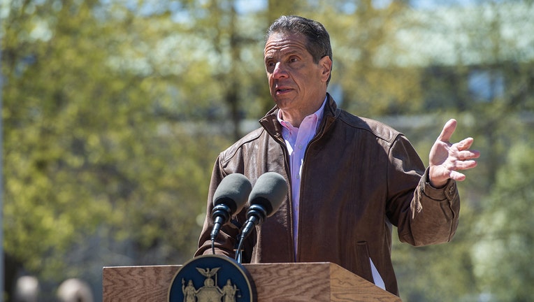 Andrew Cuomo gestures with his left hand as he speaks into microphones on a lectern; he wears a brown jacket and light-colored shirt