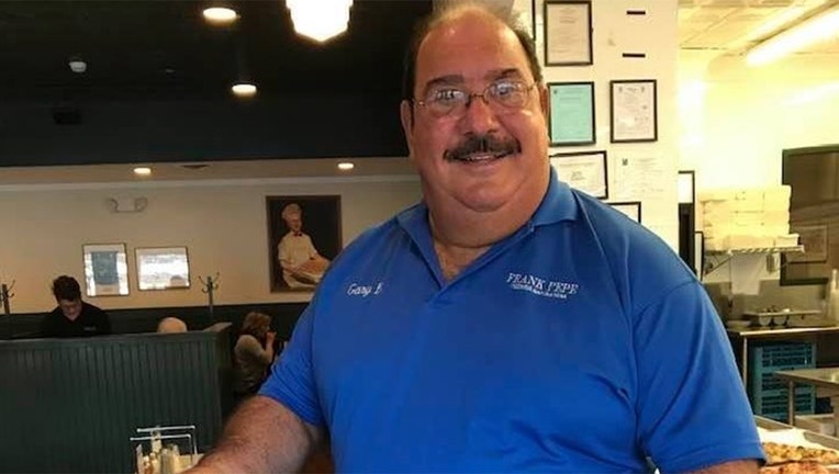 A smiling man with a mustache and glasses wearing a blue polo shirt in a pizzeria