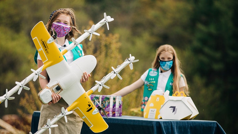 In this April 14, 2021 image provided by Wing LLC., Girl Scouts Alice Goerlich, right, and Gracie Walker pose with a Wing delivery drone in Christiansburg, Va. The company is testing drone delivery of Girl Scout cookies in the area. (Sam Dean/ Wing LLC via AP)