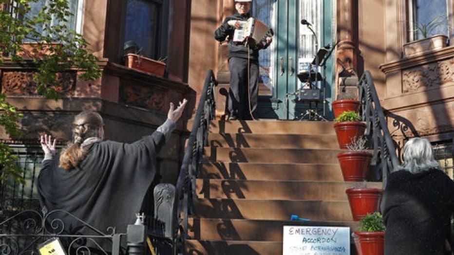 In this Wednesday, April 15, 2020 photo, choir director and neighbor Pam McAllister, left, encourages musician Paul Stein, center, to invent timely words to his songs as Stein entertains neighbors with an accordion concert from his stoop in front of his Sunset Park, Brooklyn home during the coronavirus outbreak in New York.