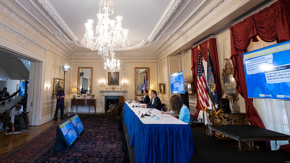Gov. Cuomo and aides sit behind a long table in a large room in the governor's mansion; an ornate chandelier hangs from the ceiling; US and New York flags are against the right wall
