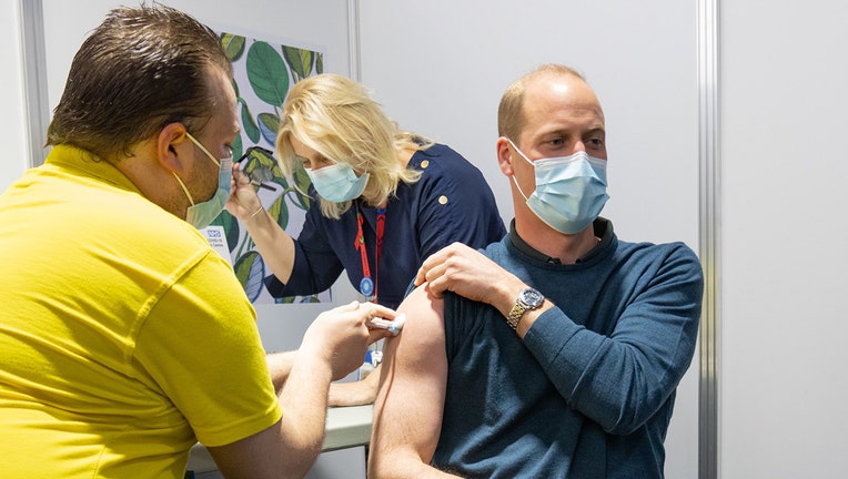 A medical worker gives a vaccine injection to Prince William while another medical worker stands behind the prince; Prince William wears a light blue mask and a bluw long-sleeved shirt