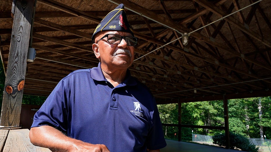 Army veteran wearing a garrison cap and navy blue polo stands under a wooden patio roof