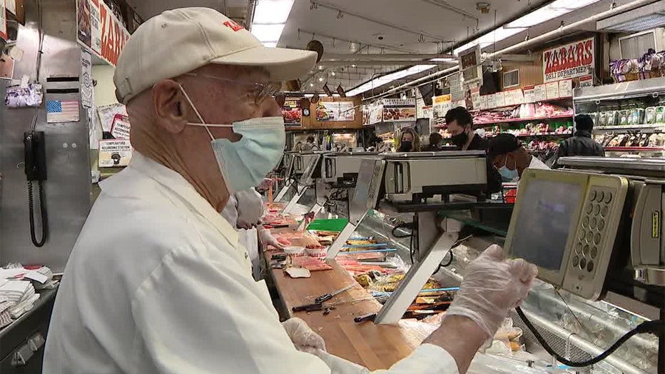 A man wearing a white hat and outfit and blue mask uses a digital panel at a deli counter