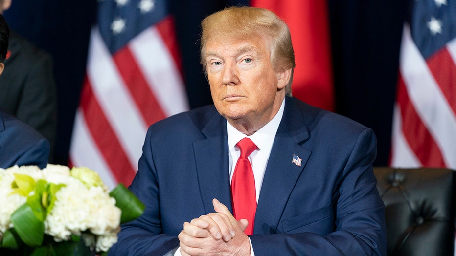 Donald Trump in a blue suit and red tie sits at a table with white flowers on the table and American flags behind him