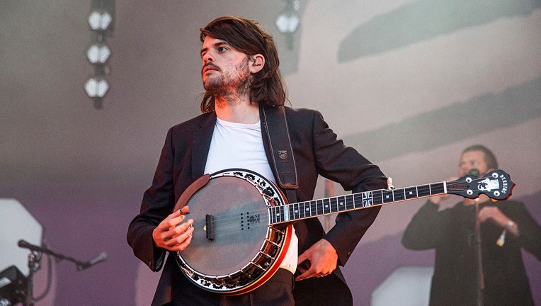 Musician with long brown hair and beard wearing a white shirt and dark jacket holds a banjo on stage