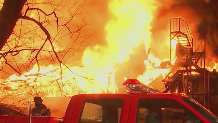 Bright orange flames engulf a residence at night; the top of a fire vehicle and a firefighter are seen in the foreground