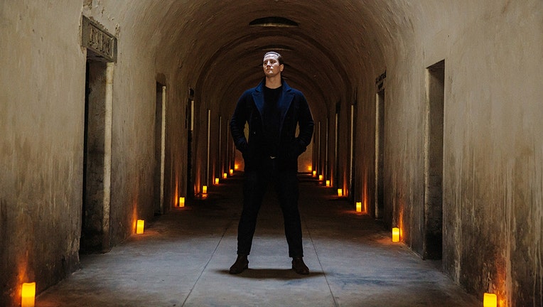 A man in a dark suit stands in a long chamber of catacombs; candles line the chamber