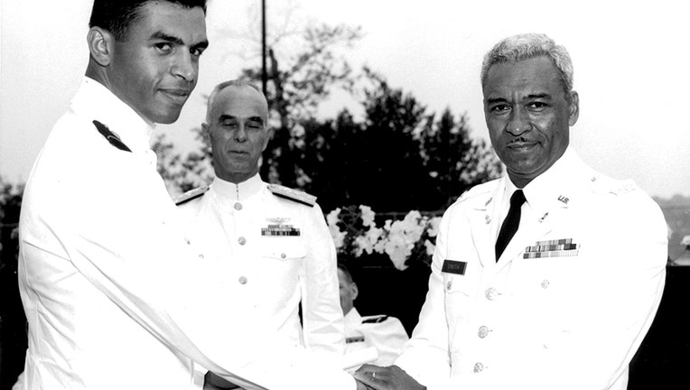 Coast Guard Academy graduate in white uniform shakes hands with his father, an Army colonel in a white uniform, as a Coast Guard admiral stands behind them