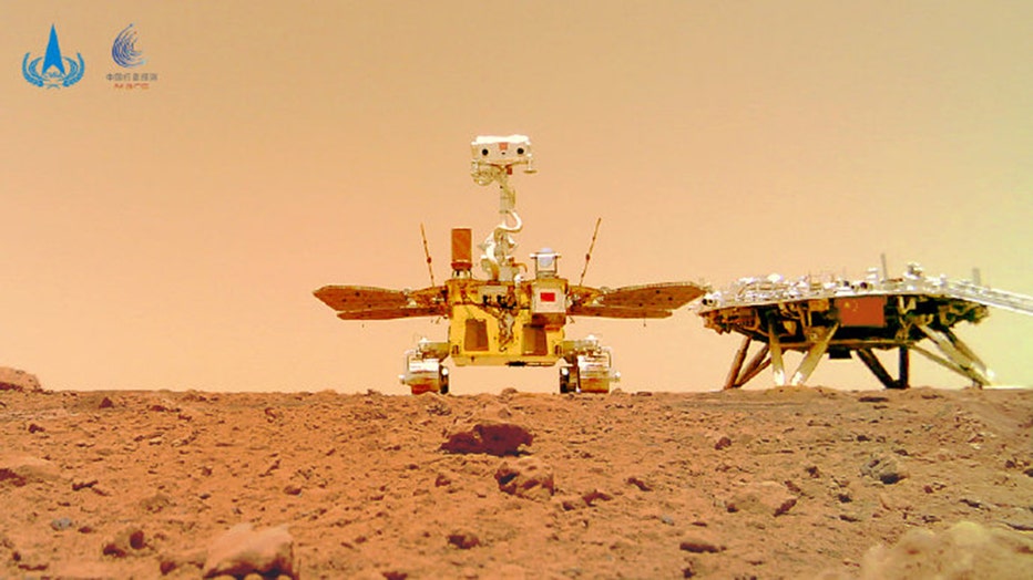 A rover and a landing platform stand on the rocky and dustry surface of Mars