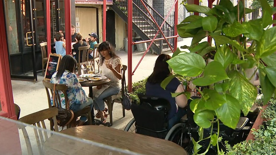 Several patrons sit at tables in an outdoor seating area along a sidewalk in East Harlem; one patron in a wheelchair is seen from behind; a pothos plant hangs in the foreground