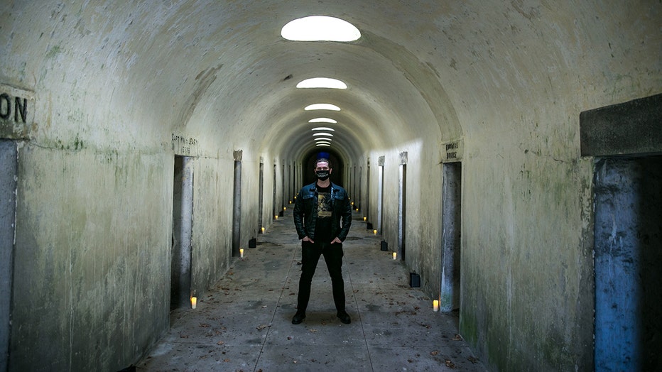 A man wearing a face mask stands in a long chamber of catacombs; candles line the chamber