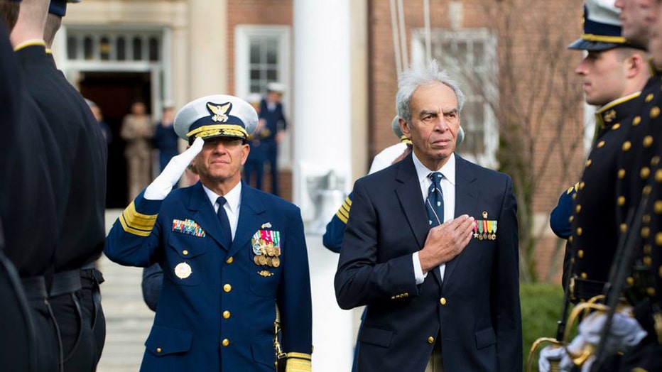 A Coast Guard officer in a blue uniform salutes as a man in a blue suit wearing medals puts his hand over his heart as other Coast Guard personnel stand nearby
