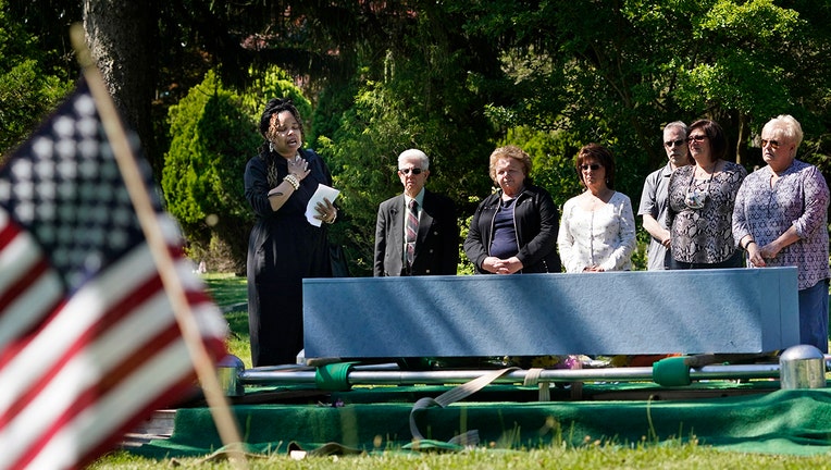 A woman holds her hand on her chest as she speaks over a casket during a burial at a cemetery; several people stand nearby; green trees and bushes are in the background; an American flag is in the foreground on the left side