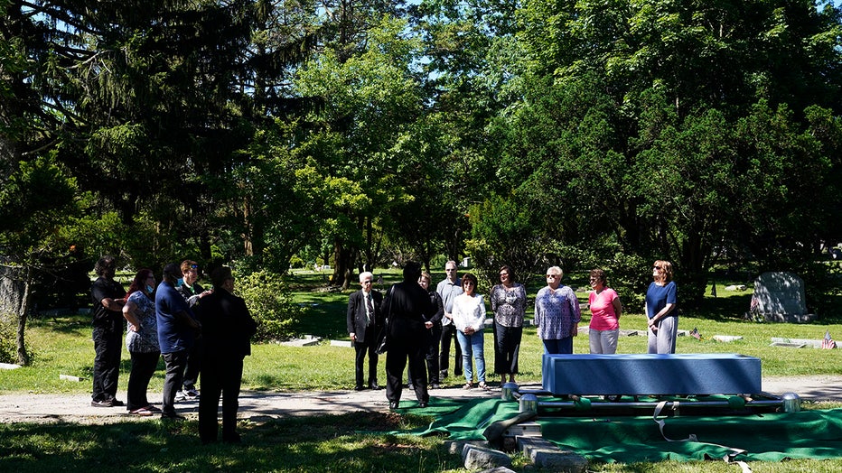 12 people stand near a casket in a cemetery; tall trees are in the background