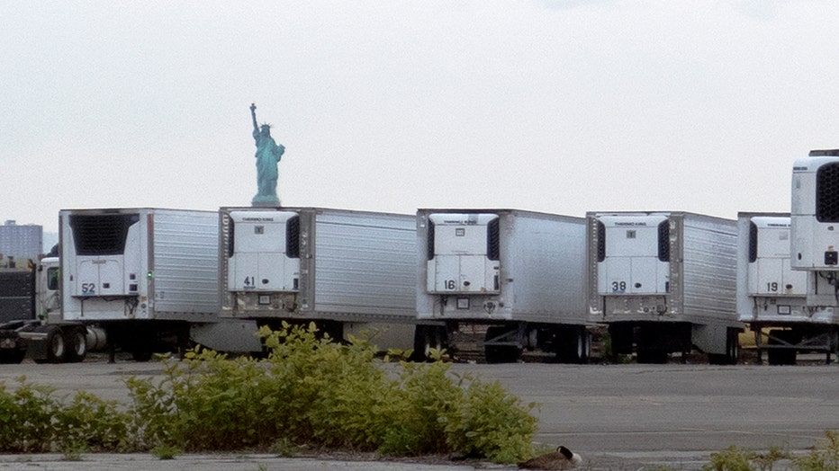 The Statue of Liberty is seen in the distance behind a row of white refrigerated trucks on a pier in Brooklyn